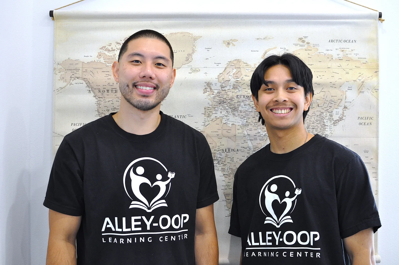 Two Alley-Oop Learning Center educators in branded shirts standing in front of a world map in the Beacon Hill center